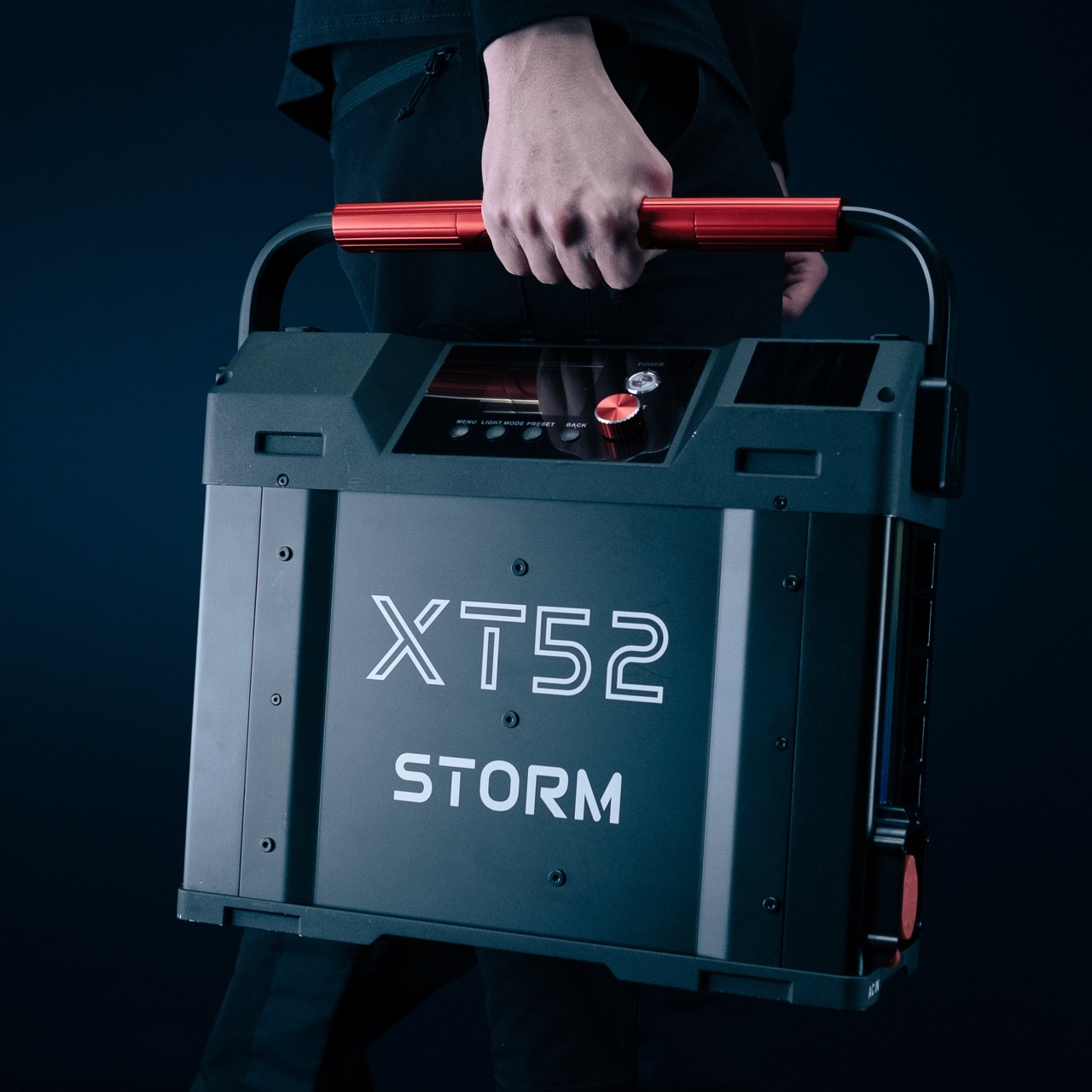 A person holding a black box with a red handle with buttons on the top. The box says “XT52 Storm” on the side