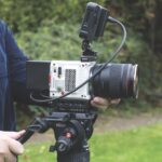 Side-view of a camera on a stand operating outside. Someone stands holding the camera.