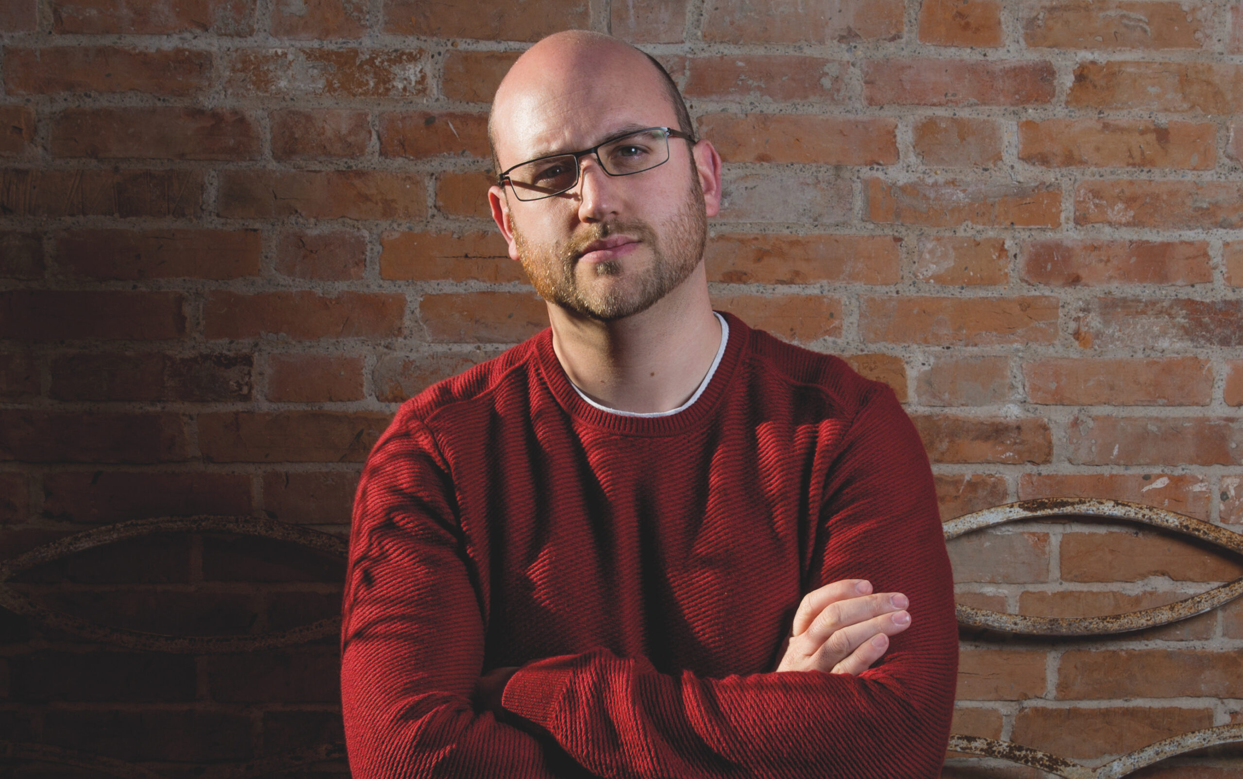 A man wearing a red jumper and glasses, looking at the camera with his arms crossed