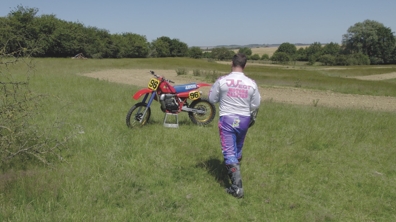 Someone in purple trousers walking towards a red motorcycle in a field