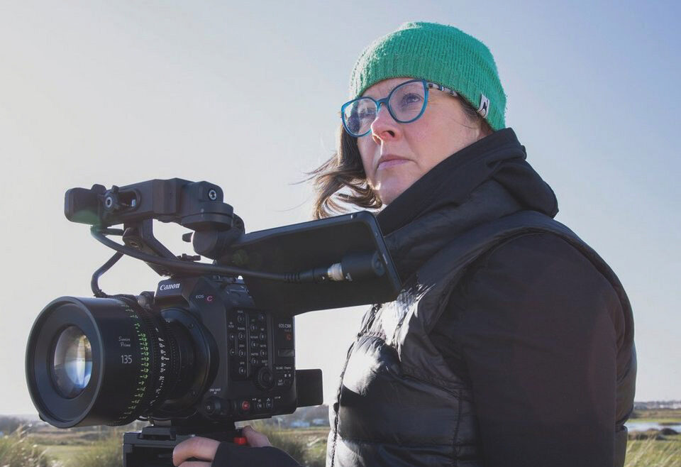 A woman wearing a black coat and green beanie, operating a large camera on a stand outside