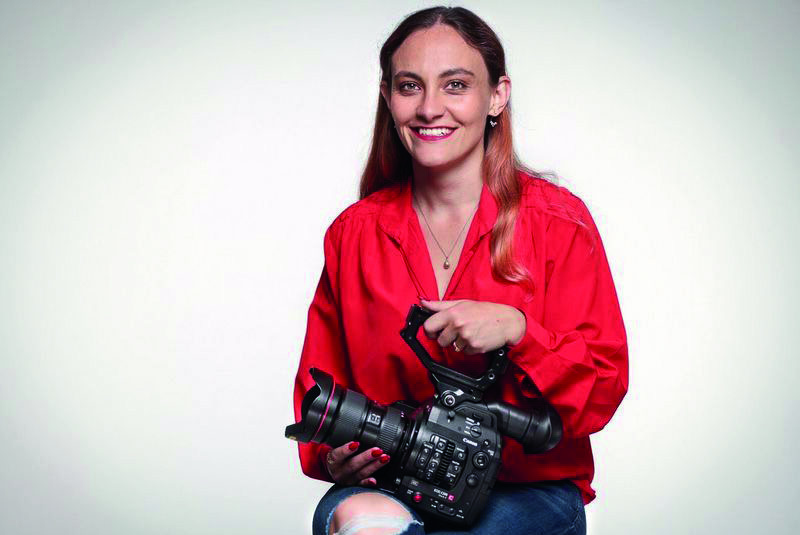 A woman sitting and smiling at the camera. She wears a red top and holds a camera on her lap.